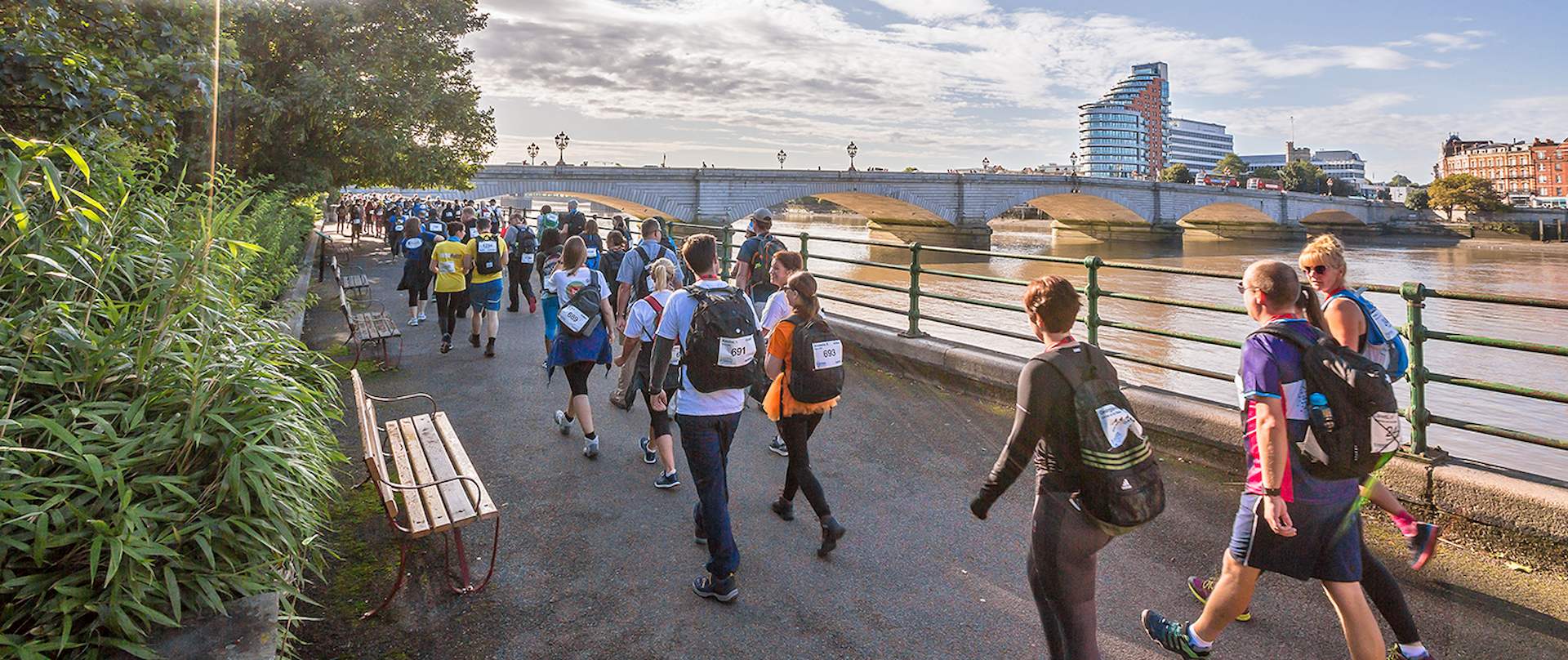 A large group of walkers walk beside the River Thames towards a bridge in the distance as part of the Thames Bridge Trek.
