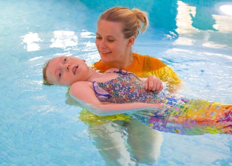 a child wearing a mermaid tail in the pool at Chestnut. She is being supported by a member of staff wearing a yellow Chestnut t-shirt