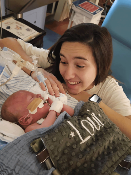 Zoe crouching next to a hospital bed, smiling at baby Loui