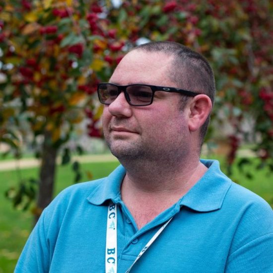 Housekeeping assistant Martin is pictured in the gardens at Chestnut Tree House, in front of an ornamental cherry tree. He wears dark glasses and a blue shirt.