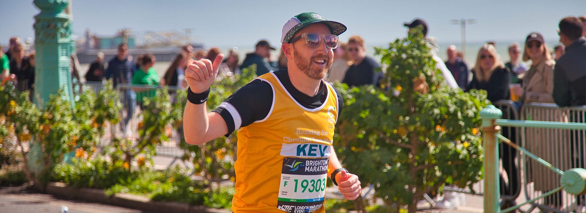 Runner taking part in a running event in Brighton. He is wearing a Chestnut Tree House running vest, smiling and giving the thumbs up while he runs along the seafront with spectators in the background