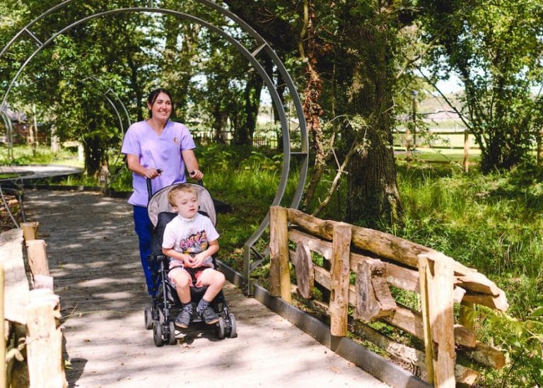 a member of the care team pushing a boy in a buggy on a board walk through our woodland walk