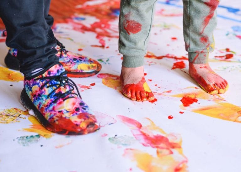 a barefoot child and adult in boots with paint on their feet standing on a large piece of paper