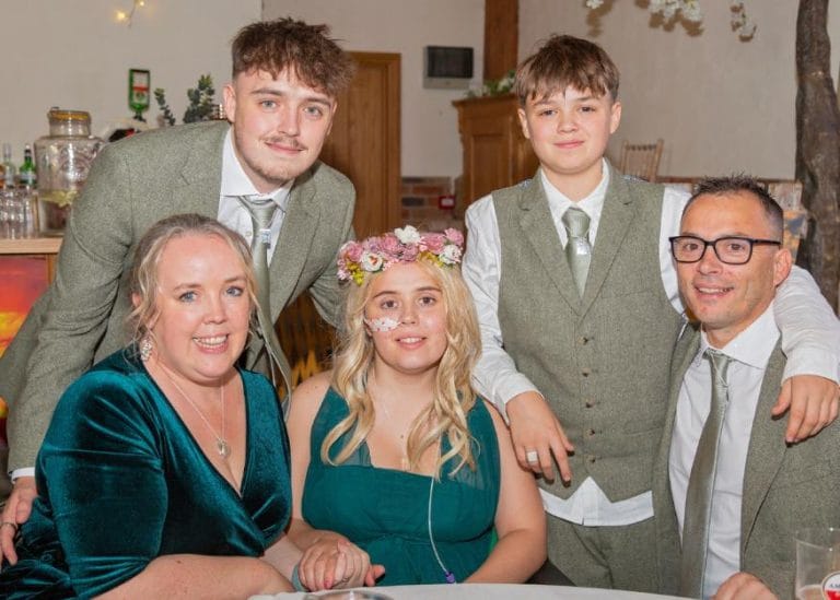 Millie and her family dressed in smart clothes. She is wearing a blonde wig and a flower crown