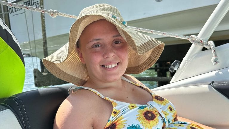 Millie on a boat, smiling at the camera. She is wearing a large sun hat and a vest top covered in sunflowers