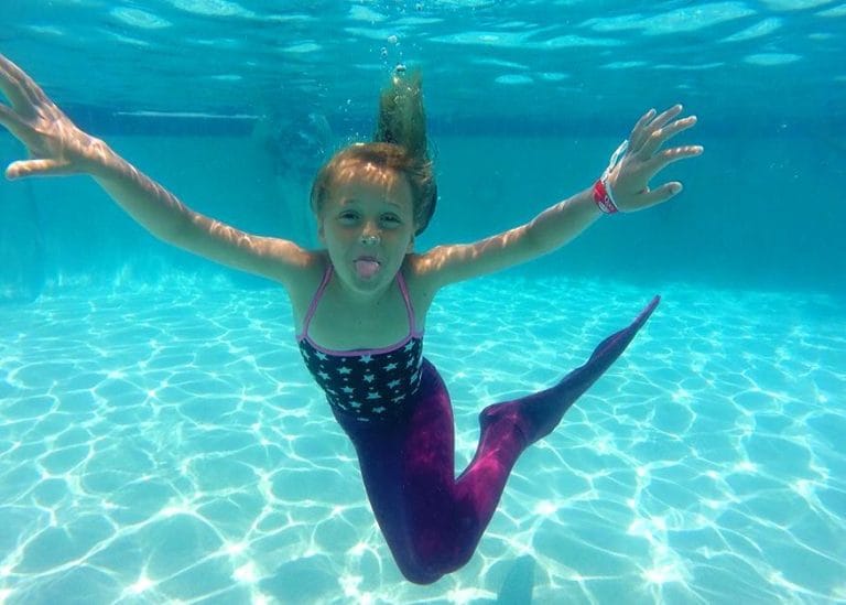 millie underwater in a swimming pool. She is dressed in a purple mermaid tail and sticking her tongue out at the camera