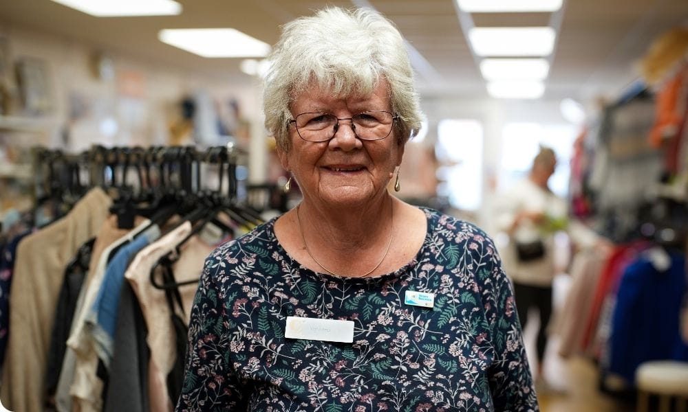 A woman stands inside a charity shop smiling.