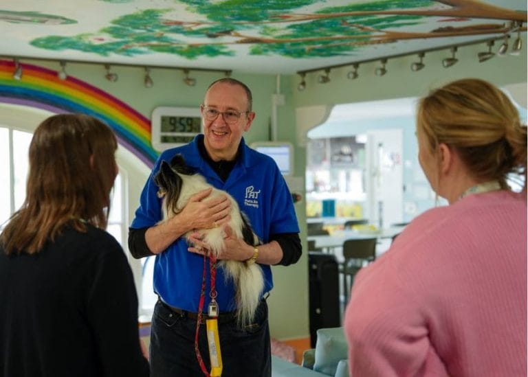 Rob, a pets as therapy volunteer holding tuppence, a small furry dog. He is talking to members of staff at Chestnut Tree House