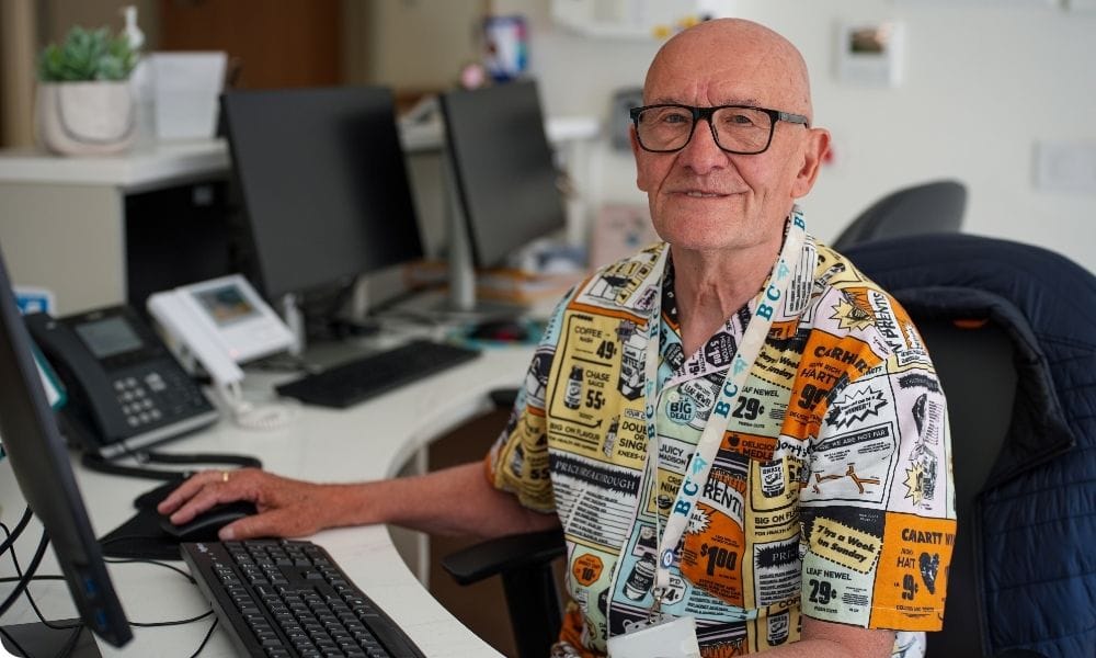 A man sits at a reception desk wearing a patterned shirt and operating a computer