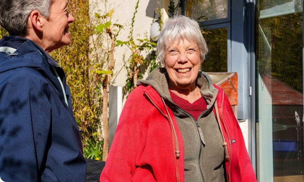 A woman in a red jacket stands outside smiling in the sunshine