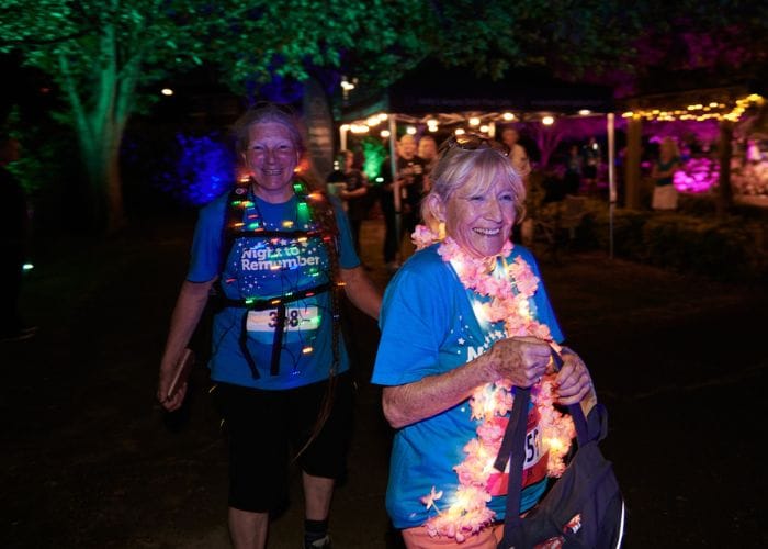 two participants at night to remember, walking through a dark area with trees illuminated and festoon lighting. They are wearing a lei and light up necklace and smiling