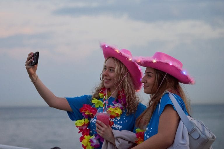 two participants in night to remember wearing pink cowboy hats and leis taking a selfie in front of the sea